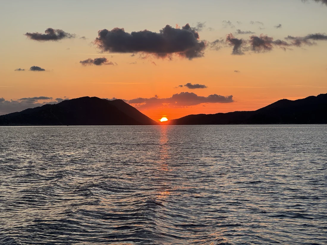 Die orangefarbene Sonne versinkt zwischen dunklen Küstenhügeln im Meer.